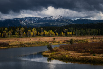 Fototapeta premium Breathtaking landscape featuring a winding river, golden fall foliage, and a snow-capped mountain under dramatic storm clouds. Perfect for travel and environmental themes.