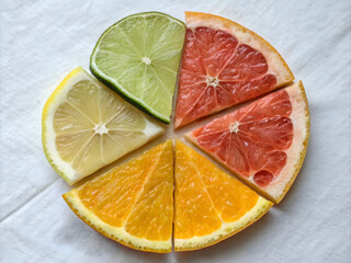 Top view of assorted citrus fruit segments including ruby red grapefruit orange and yellow lemon wedges arranged in circular composition on white textured fabric background
