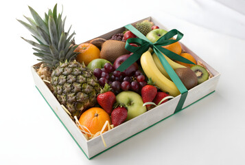 Large rectangular gift box brimming with whole pineapple bananas strawberries apples grapes and oranges tied with elegant green satin ribbon bow on clean white background
