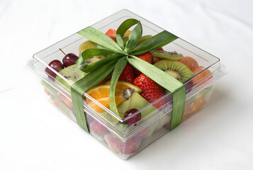 Transparent plastic gift container filled with colorful sliced kiwi strawberries blueberries raspberries oranges tied by decorative green textured ribbon bow on white background