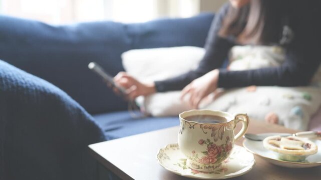  Relaxing morning scene or afternoon tea with teacup,sweet pie on coffee table,Blurry woman use smartphone resting on sofa in background,Concept slow life,mental health,quiet moment of wellness at hom