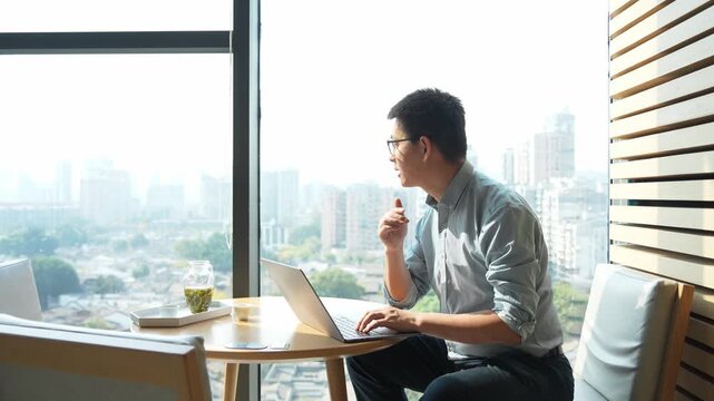 Young man working on a laptop by the window while gazing at the cityscape