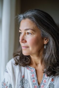 Middle-aged woman with long gray hair wearing a white embroidered blouse gazes thoughtfully out of a window, soft natural light illuminating her face
