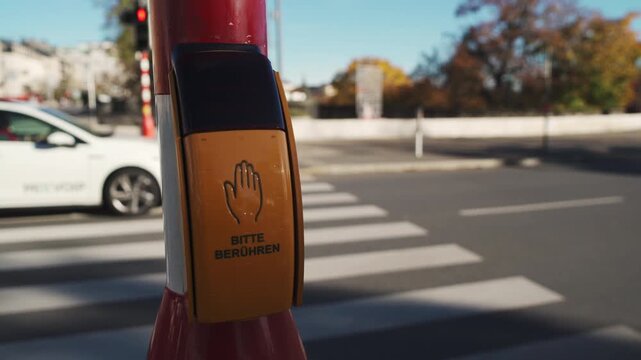 A yellow pedestrian crossing button with the word bitte beruhren is mounted on a pole near a street intersection, with blurred cars and trees in the background. 