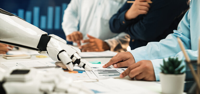 A robot hand interacts with human colleagues during a business meeting. They analyze various charts and graphs, showcasing collaboration in a modern office setting.Noogenesis