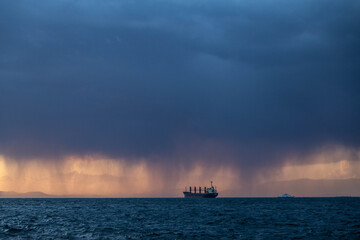 Obraz premium Dramatic sunset over the Aegean Sea in Izmir, Turkey. Large cargo ship on the horizon during a rain shower with dark heavy clouds and golden light on a windy day in January.