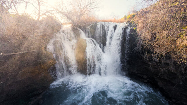 A hiker viewed from behind on a wooden walkway, observing the misty Pozo Redondo waterfall in Calmarza, enjoying a moment of solitude and connection with the powerful nature of the Aragon region.