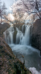 Artistic long exposure of the Pozo Redondo waterfall in Calmarza during winter, with the water flowing like silk over the rocks and framed by the bare branches of surrounding trees against a cloudy 