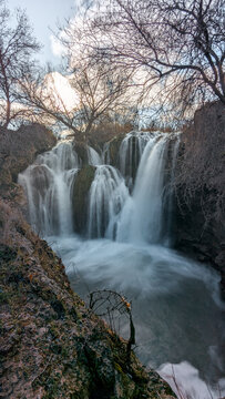 Artistic long exposure of the Pozo Redondo waterfall in Calmarza during winter, with the water flowing like silk over the rocks and framed by the bare branches of surrounding trees against a cloudy 