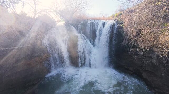 The stunning Pozo Redondo waterfall in Calmarza, Zaragoza, captured with a bright sun flare piercing through the mist, highlighting the beauty of the cascading water over the rugged rock formations.