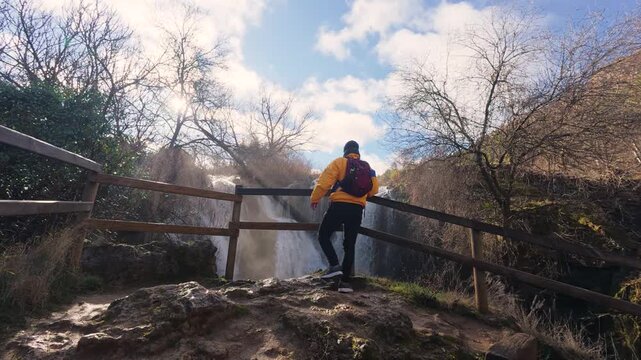 A lone hiker contemplates the impressive Pozo Redondo waterfall from a wooden footbridge viewpoint in Calmarza, enjoying the natural scenery and the power of the cascading water in Zaragoza.