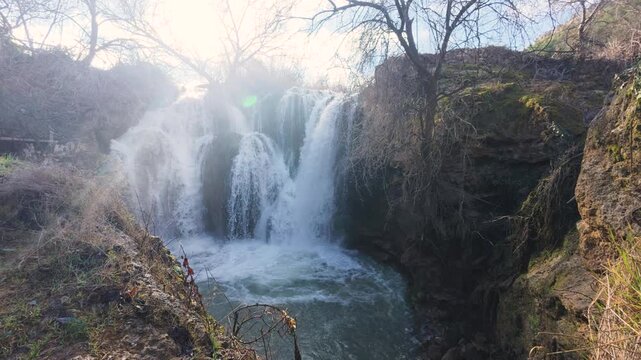 Majestic view of the Pozo Redondo waterfall in Calmarza, Zaragoza, with powerful streams of water cascading over rocks and a beautiful sun flare illuminating the mist on a bright sunny day.