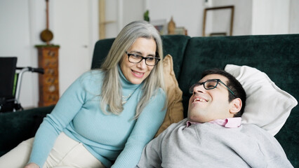 Senior mother spending quality time by her adult son with a physical disability on a couch at home,...