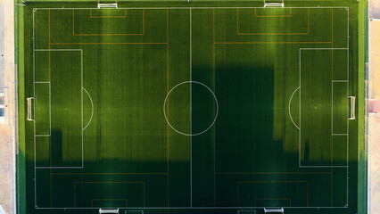 Green artificial turf soccer field with white markings and goalposts, seen from an aerial perspective with long shadows © DMegias