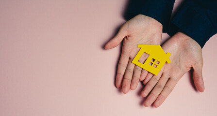 Buying a new house concept. Person holds yellow house shaped cutout their hands, symbolizing home ownership and real estate. soft pink background adds warm and inviting feel