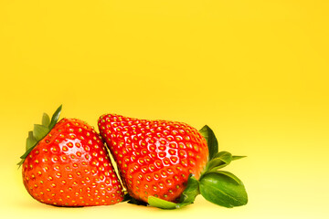 Two ripe red strawberries with green leaves positioned closely together on a bright yellow background, showcasing their vibrant color and texture