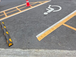 A designated disabled parking space on asphalt, marked with a white wheelchair symbol, yellow hazard lines, an orange traffic cone, and a black-and-yellow wheel stop.
