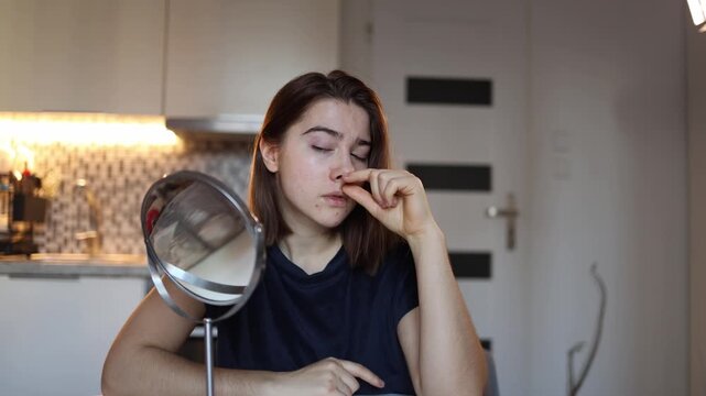 Young woman touches her nose while watching reflection unhappy with its shape thinking about surgical correction inner conflict self doubt home beauty moment