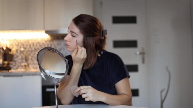 Young woman learns makeup basics applying foundation and concealer while studying her face in mirror practicing technique and hand control during calm home beauty routine