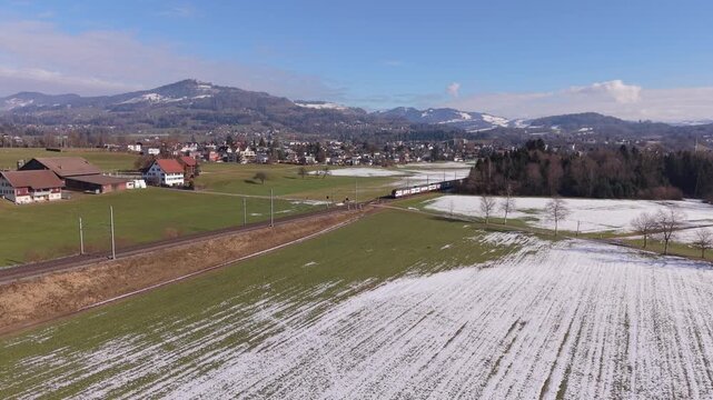 Aerial push-in shot of a passenger train traveling on railway tracks through the Swiss countryside.