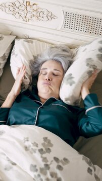 Senior woman suffering from insomnia, covering her ears with a pillow because her husband is snoring loudly in bed. Annoying sleeping problem. Vertical