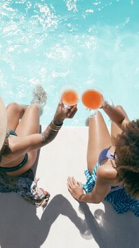 High angle view of two young women in swimsuits toasting with refreshing cocktails while relaxing with their feet in a sunny swimming pool. Vertical