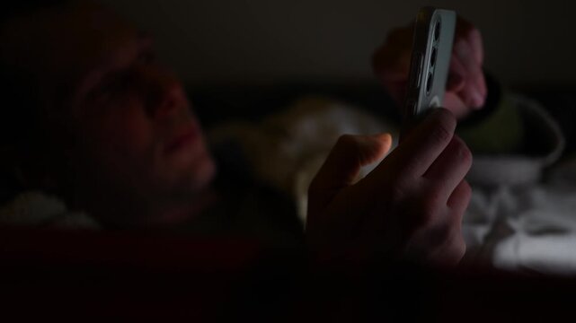 A sleepless young man lies awake in his bedroom at night, using his smartphone scrolling social media. Digital dependence and insomnia concept. 