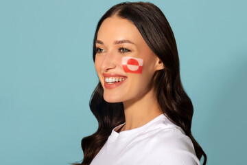Fototapeta premium National Symbol. Cheerful lady with painted flag of Greenland on her cheek smiling, showing support, posing over blue background
