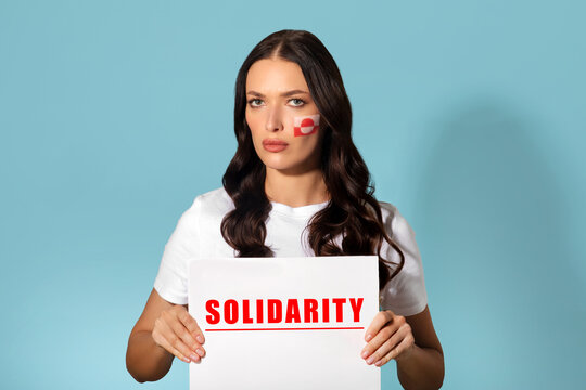 Lady holding sign saying Solidarity while showing support for issues in Greenland, posing with flag painted on cheek, blue studio background