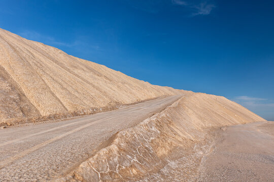 Industrial potash mine pit with terraced salt slopes and service road leading upward under deep blue sky. Arid mining landscape shows mineral textures, extraction scale, isolation, raw geology