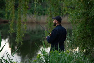 Young bearded man in a black cap standing by the river, fishing with a spinning rod, half-turned view, surrounded by lush green grass.