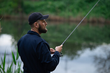 Young bearded man in a black cap standing by the river, fishing with a spinning rod, half-turned view, surrounded by lush green grass.