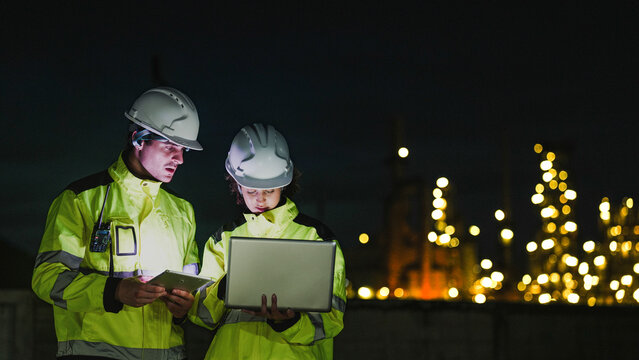 Two industrial workers in hard hats and high-vis gear work at night. They use a laptop and tablet to monitor a lit-up refinery plant during a night shift.