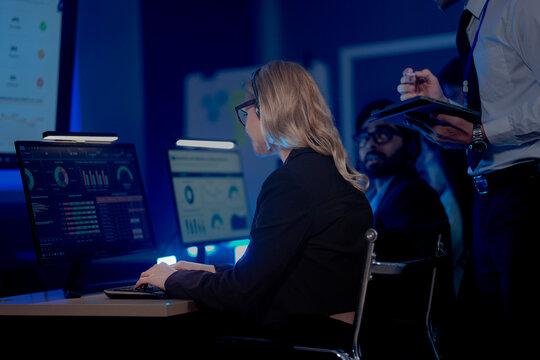 Professional female analyst monitoring business data on multiple computer screens. Young woman working with financial dashboard charts in modern blue light office room.