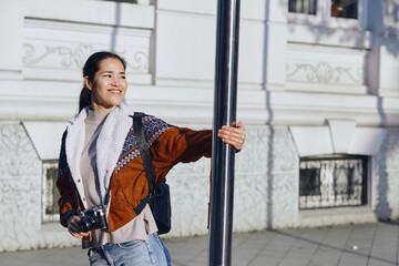 Woman with camera and backpack leaning on a pole on a sunny street, smiling, urban backdrop, casual...