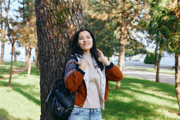 Obraz premium Woman leaning against a tree in a park, autumn sunlight and nature setting, casual sweater and backpack for outdoor lifestyle and travel imagery