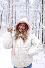 Uśmiechnięta młoda kobieta w jasnej zimowej kurtce z kapturem stoi w zaśnieżonym lesie i wskazuje palcem w górę. Zimowy krajobraz, pozytywne emocje, natura i koncepcja inspiracji lub pomysłu. © Izabela