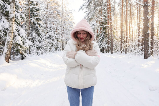 Młoda kobieta w jasnej zimowej kurtce i kapturze stoi na ośnieżonej leśnej drodze, obejmując się ramionami. Zimowy krajobraz, miękkie światło słoneczne i spokojna atmosfera. Koncepcja zimy, natury.