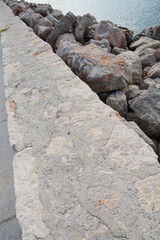 Fototapeta premium A long stone breakwater built with large grey rocks and a paved concrete path sits next to the calm blue sea water under a daylight sky
