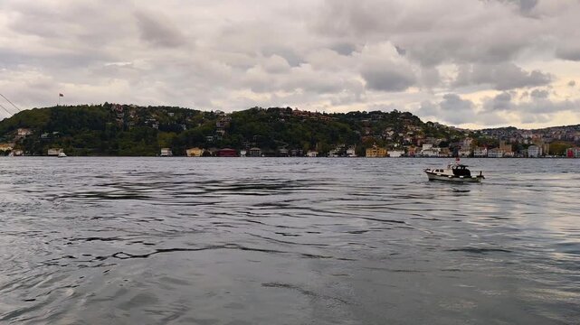 Scenic View of Luxury Yacht and Small Boat Cruising on the Bosphorus Strait with Green Hills, Traditional Waterfront Mansions (Yalı), and Cloudy Sky in Istanbul, Turkey