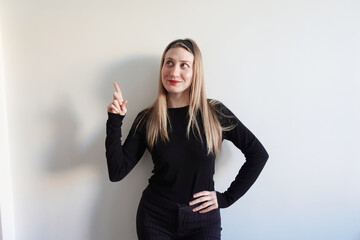 Młoda kobieta w czarnym stroju wskazuje palcem w górę i patrzy w bok na jasnym tle. Koncepcja pomysłu, inspiracji i komunikacji wizualnej. © Izabela