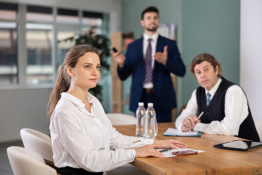 Economists or engineers employees of corporation sitting at table, talking and discussing, colleague with remote control in background, assist and helps manage presentation. Specialists during meeting