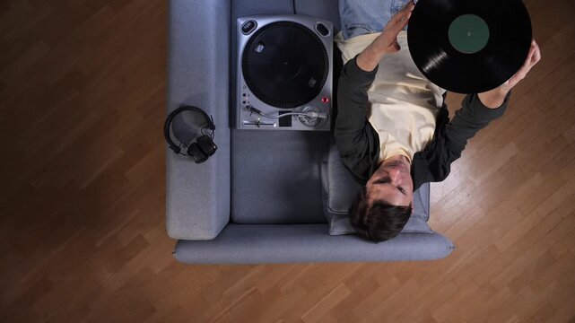 Young man lying on gray sofa holding a vinyl album, turntable and headphones beside him in a cozy living room, enjoying retro analog music and relaxing at home