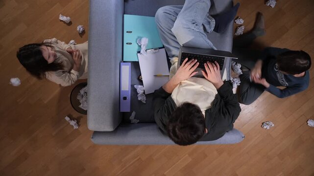 Father trying to focus on his laptop while working remotely, with his daughter throwing crumpled paper around and interrupting his concentration during the day
