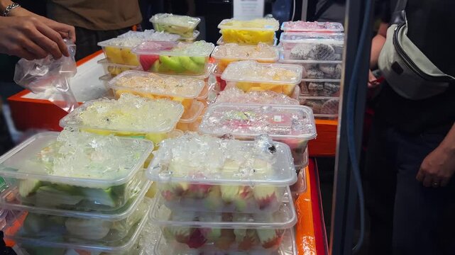 Colorful pre cut fruit boxes stacked on ice at market stall while customers point and choose. Food service, vending, freshness, and ready to eat fruit salad concept.