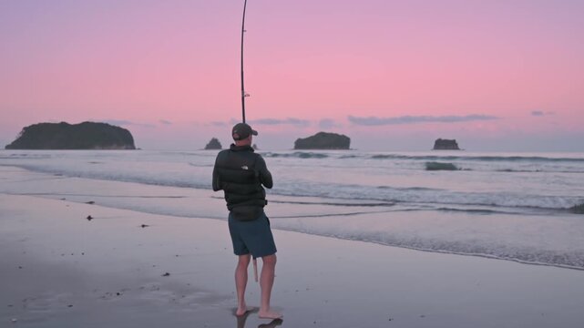 A man fishes at dawn on Whangamata, Coromandel Peninsula, New Zealand, New Zealand. He is surfcasting, hoping to catch fish in the surf. The pink sky reflects on the wet sand.