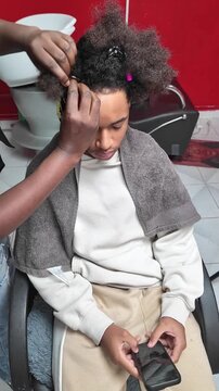 Teen boy sitting in salon chair looking at phone while stylist braids the front of his afro hair