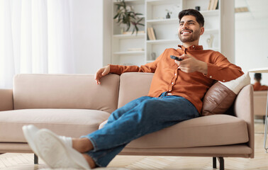 Obraz premium Portrait of Arab guy watching TV show or film, holding remote control, switching channels. Young smiling man spending weekend free time sitting on comfortable sofa at home in living room, copy space