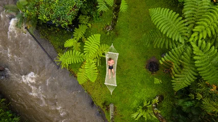 Fotobehang Bos rivier Aerial top down view of a person relaxing in a hammock beside a flowing river surrounded by lush green tropical plants and forest.  © Bpato Media