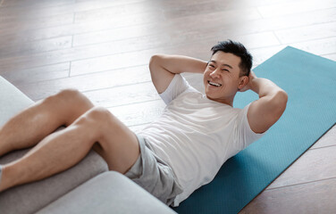 Fototapeta premium Sporty asian mature man exercising abs muscles on yoga mat, putting legs on couch and smiling at camera. Active sportsman leading active lifestyle, working out at home
